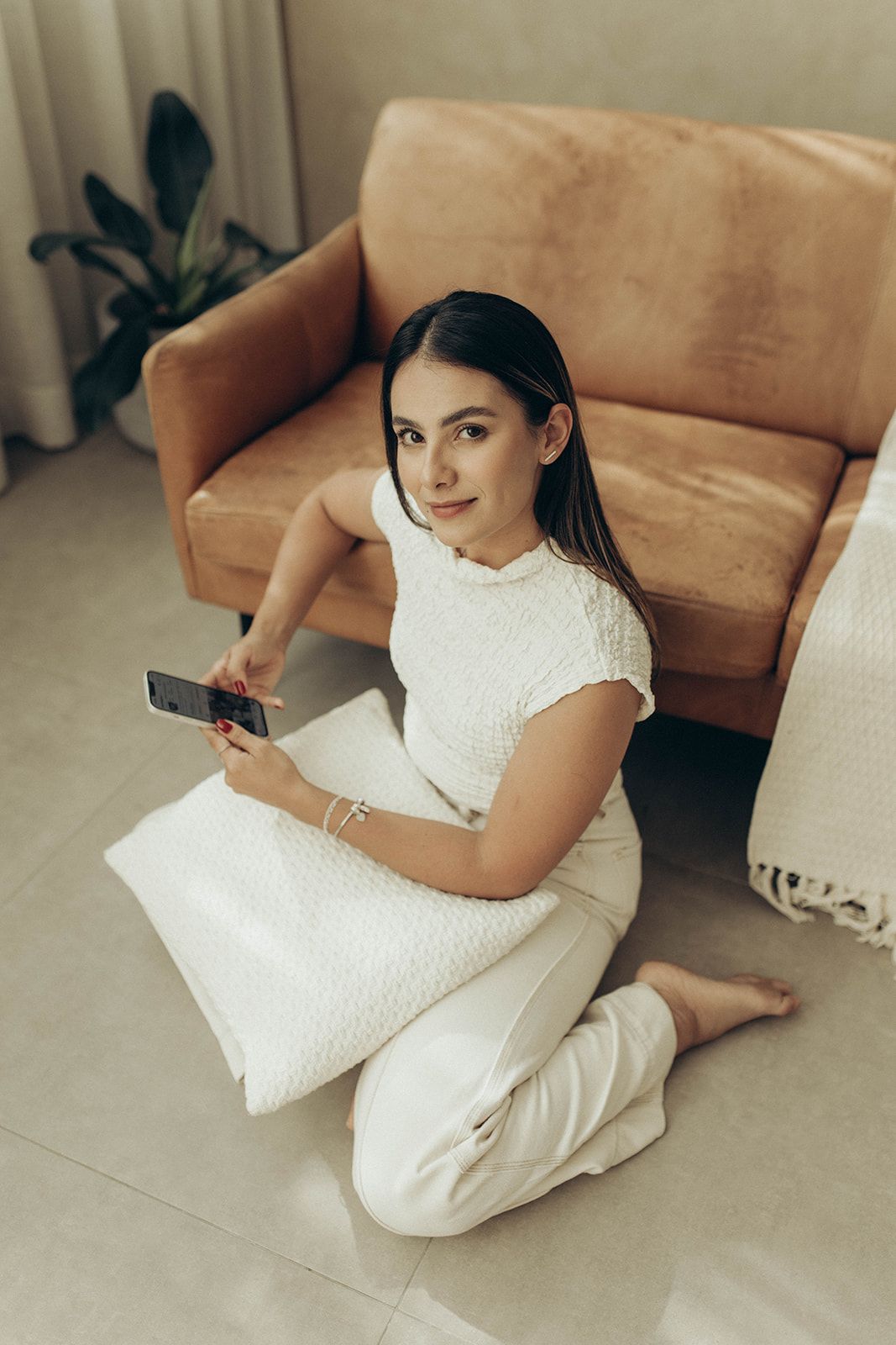 Leticia Gabriela Inacio sentada no chão, com um celular, em frente a um sofá.